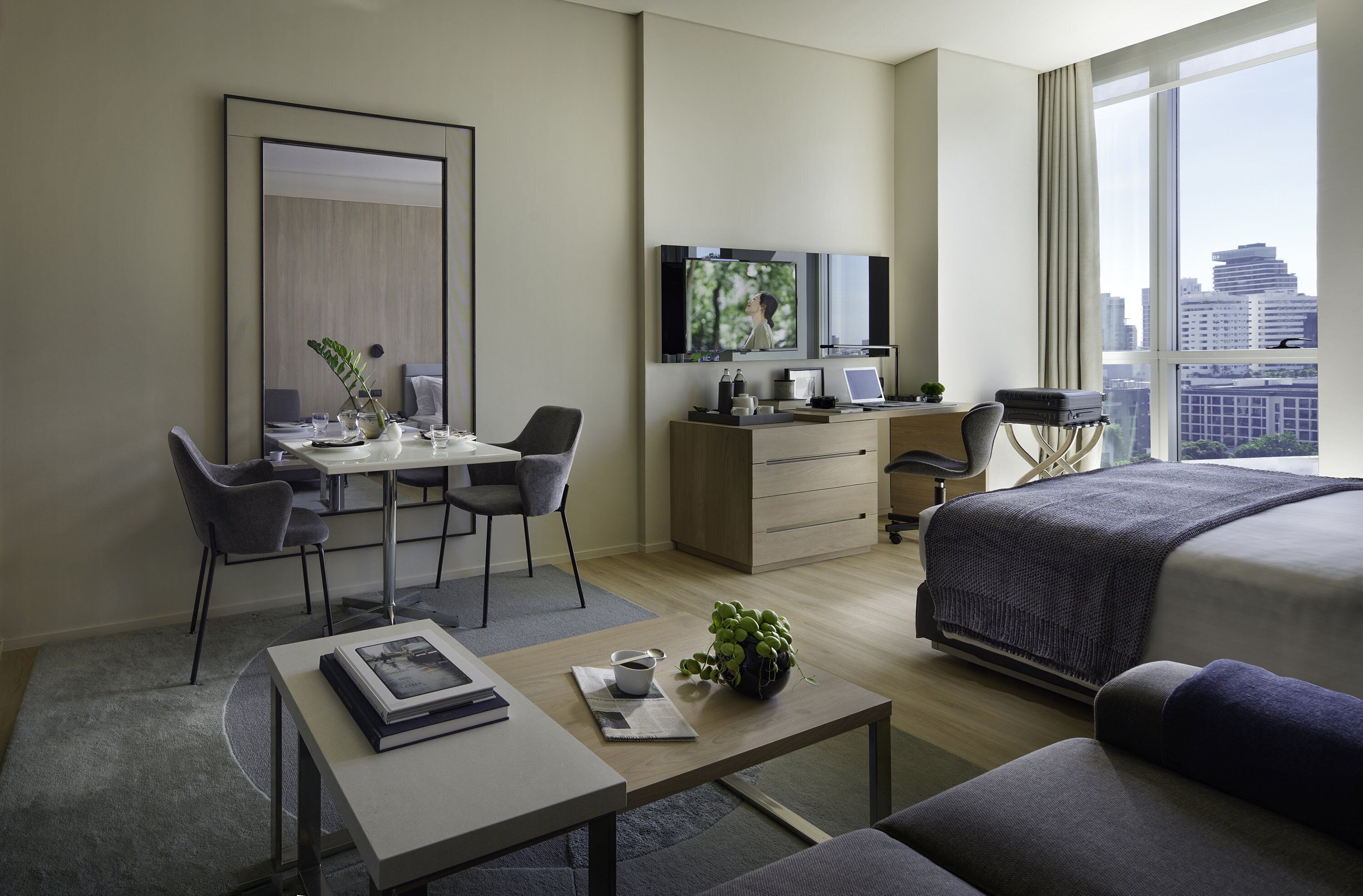 A modern hotel room with a bed, a desk with a computer, a TV, and a large window with a city view.