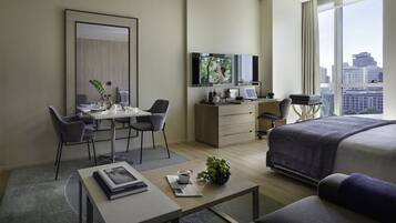 A modern hotel room with a bed, a desk with a computer, a TV, and a large window with a city view.