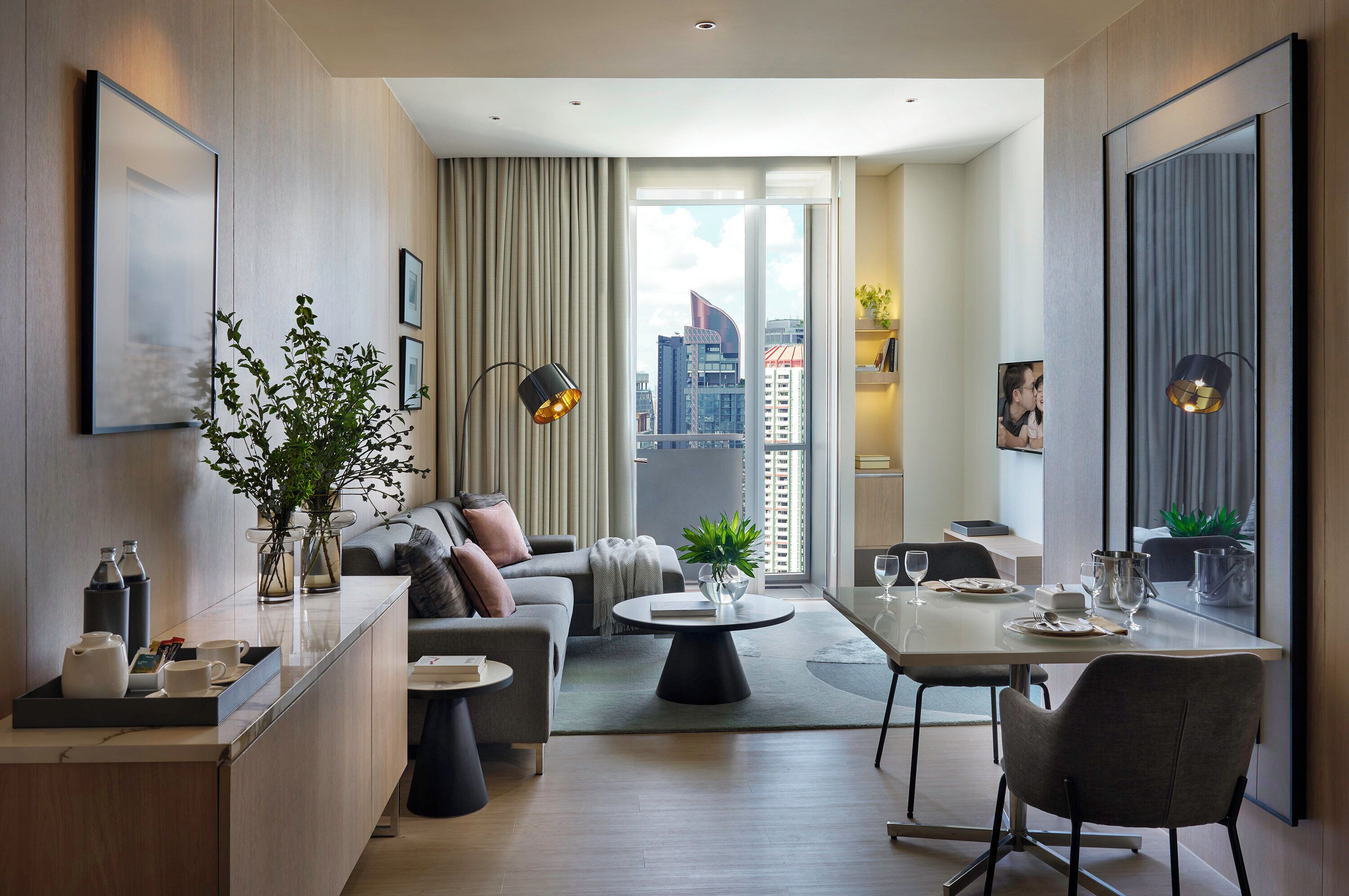 A modern hotel room with a sofa, a dining table, and a view of the cityscape.