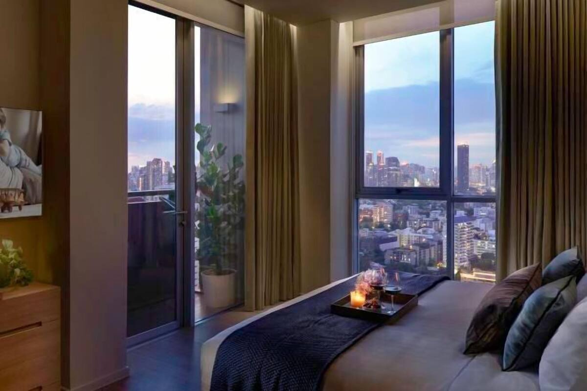 A modern bedroom with a large bed, a nightstand, a potted plant, and a city view through large windows.