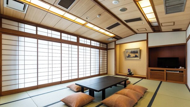 Une pièce de style japonais traditionnel, avec un sol recouvert de tatamis, une table basse, des coussins, un téléviseur et un tableau accroché au mur.