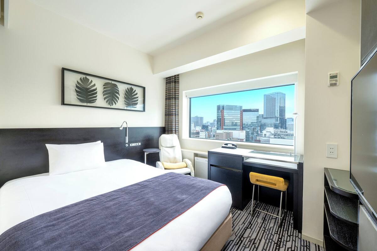 A hotel room with a large bed, a desk, a chair, a TV, and a window with a city view.