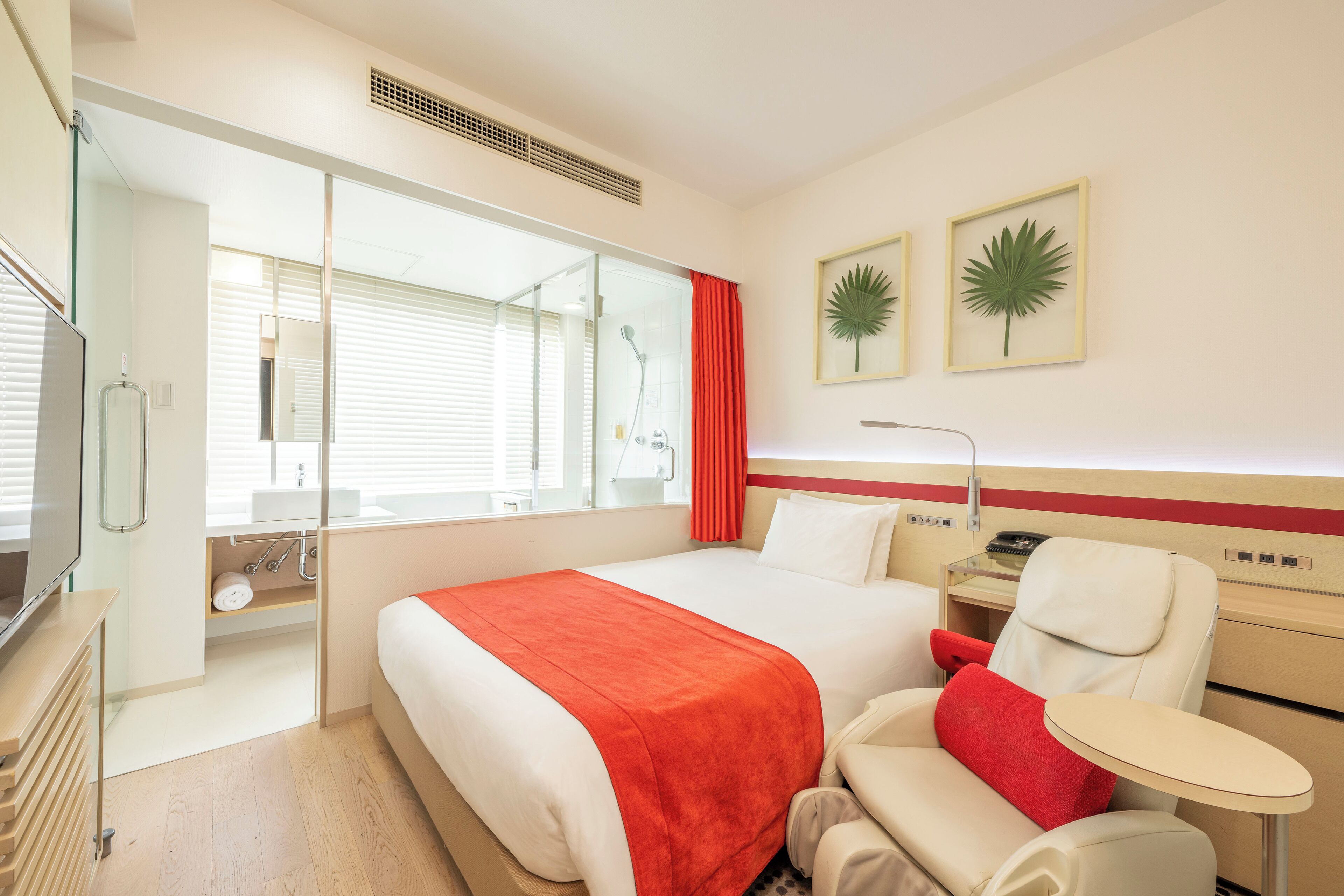 Quarto de hotel com cama, escrivaninha, cadeira, mesa pequena, televisão e janela com persianas.