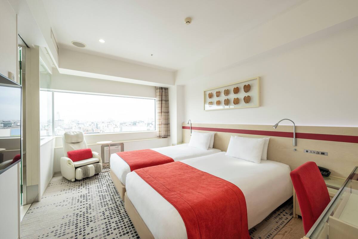 A hotel room with two beds, a red chair, a small table, and a window with a city view.