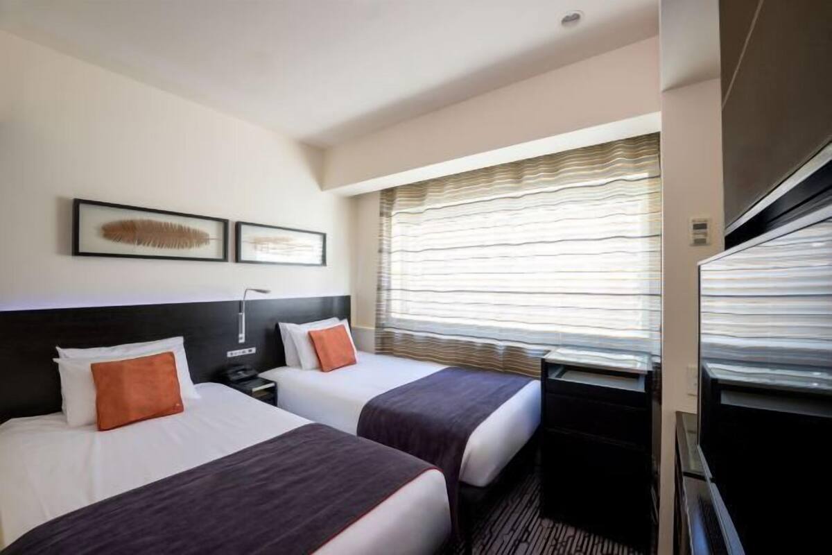 A hotel room with two beds, a desk, a TV, and a window with blinds.
