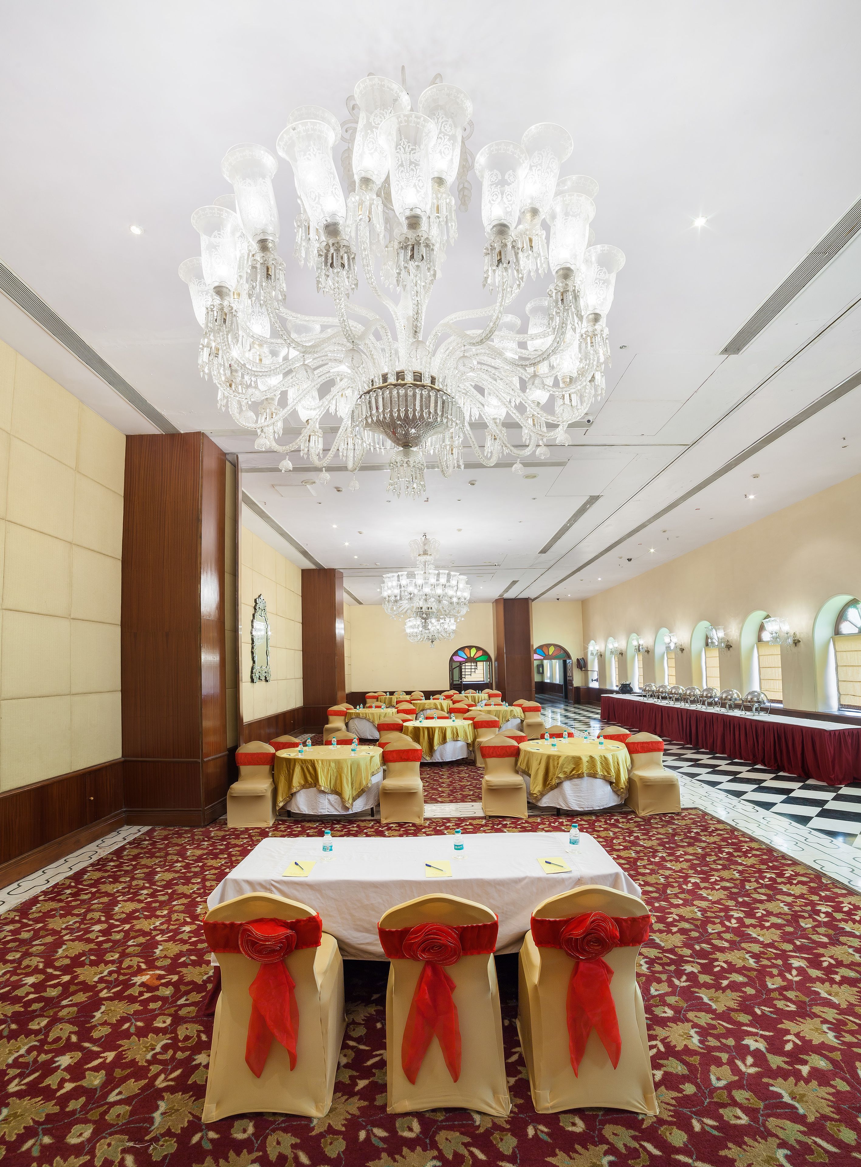 A banquet hall with a large chandelier, round tables with red sashes, and a patterned carpet.