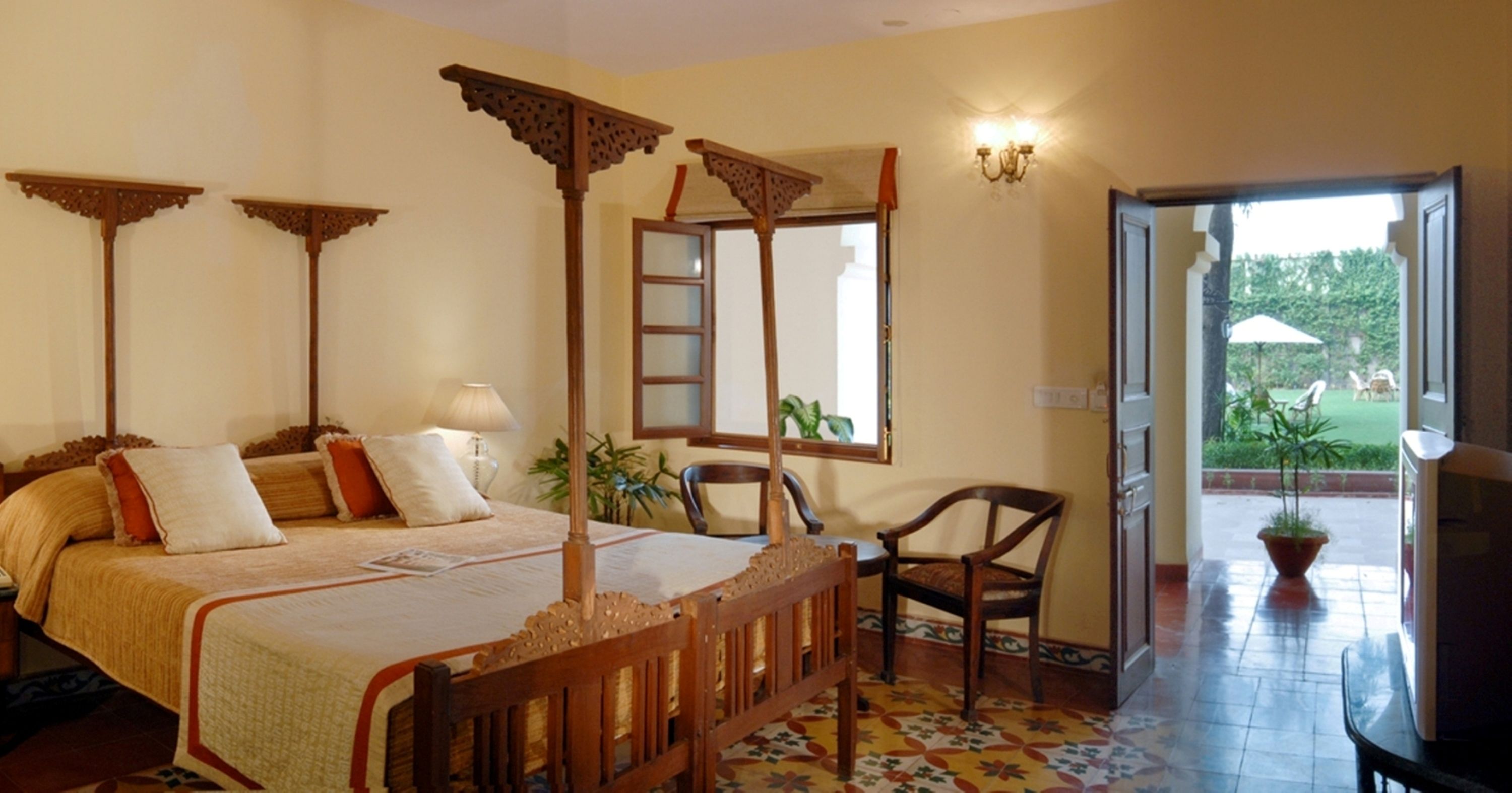 A bedroom with a four-poster bed, a chair, a desk, and a view of the garden through an open door.