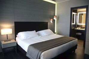 Una habitación de hotel moderna con una cama grande, una mesita de noche con lámpara, y un baño visible a través de una puerta abierta.