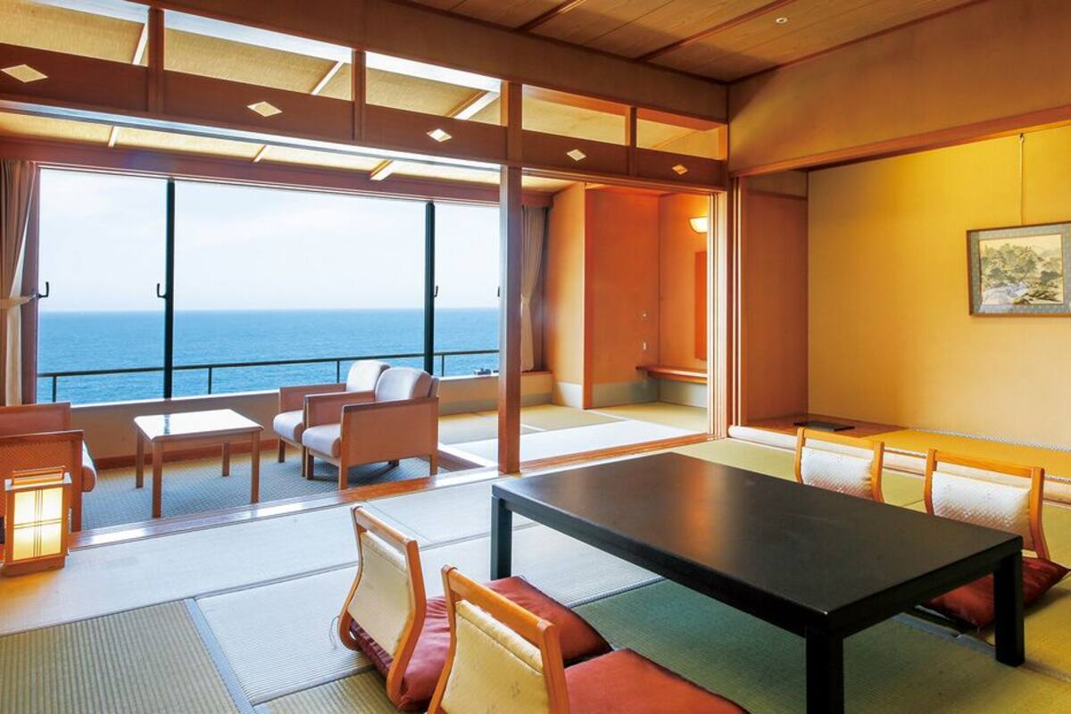 A room with tatami mats, a low table, chairs, a view of the sea, and a painting on the wall.