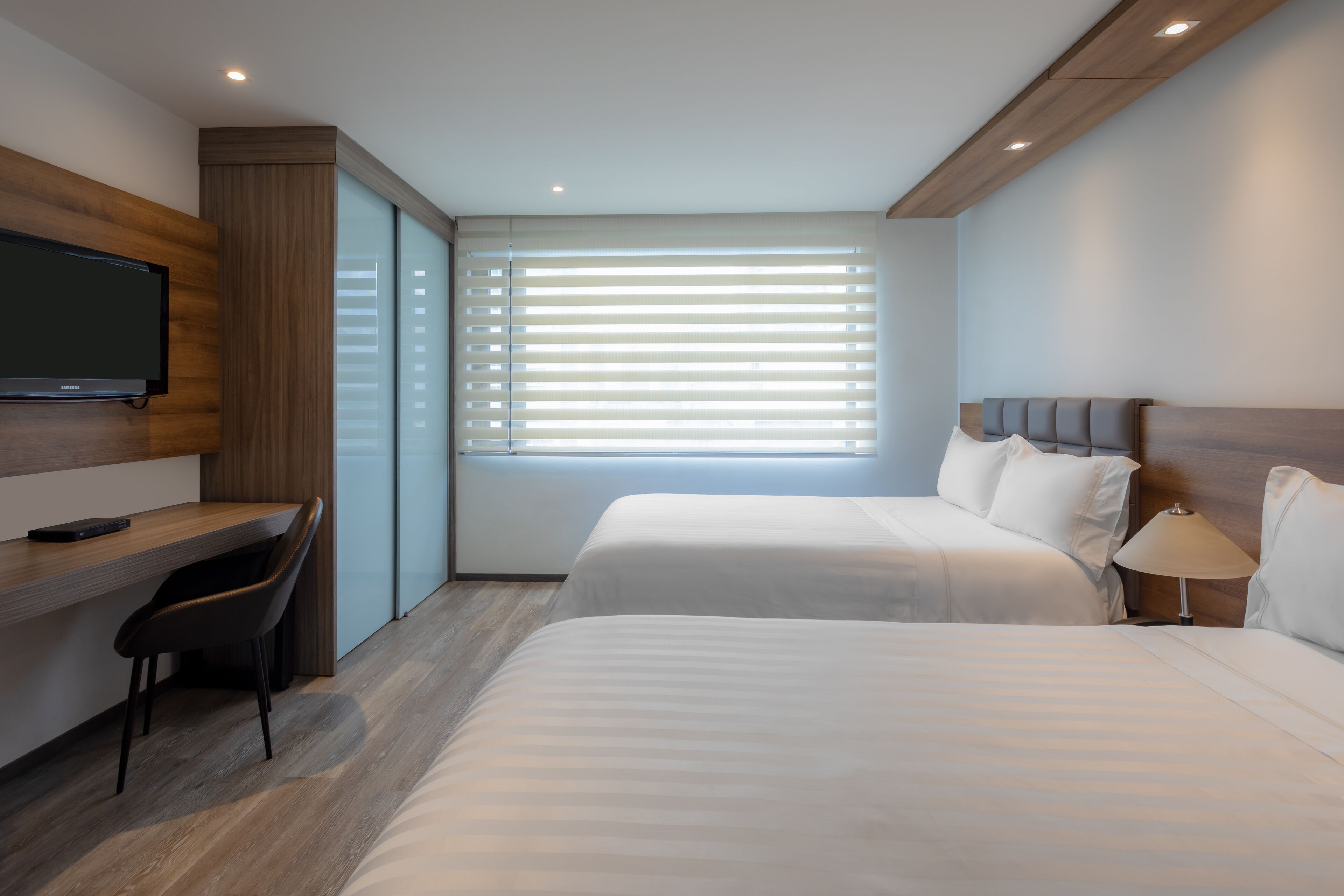 A modern hotel room with a wooden desk, a flat-screen TV, two beds, and a large window with blinds.