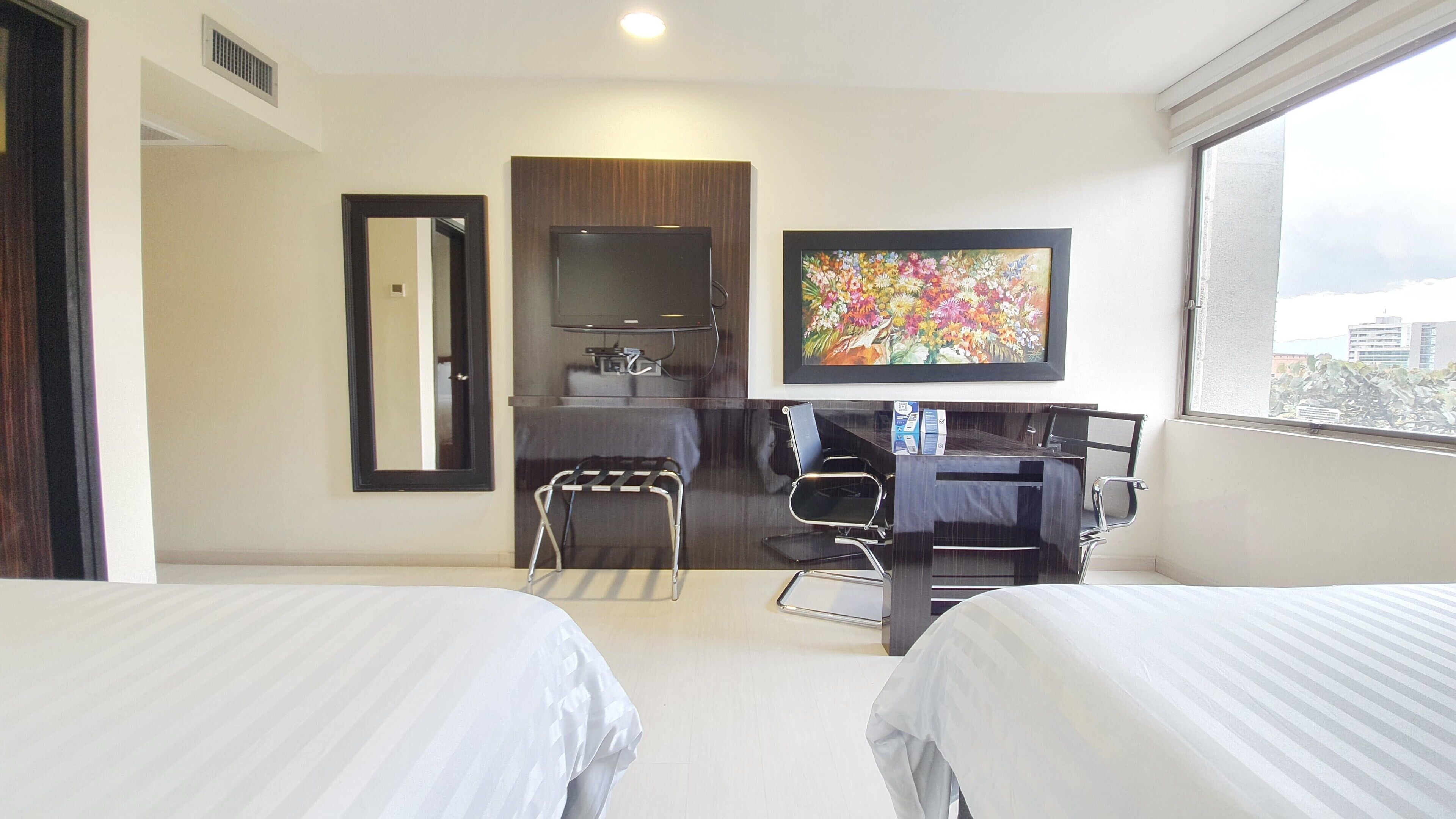 Habitación de hotel con dos camas, un escritorio con una silla, un televisor y un cuadro en la pared.