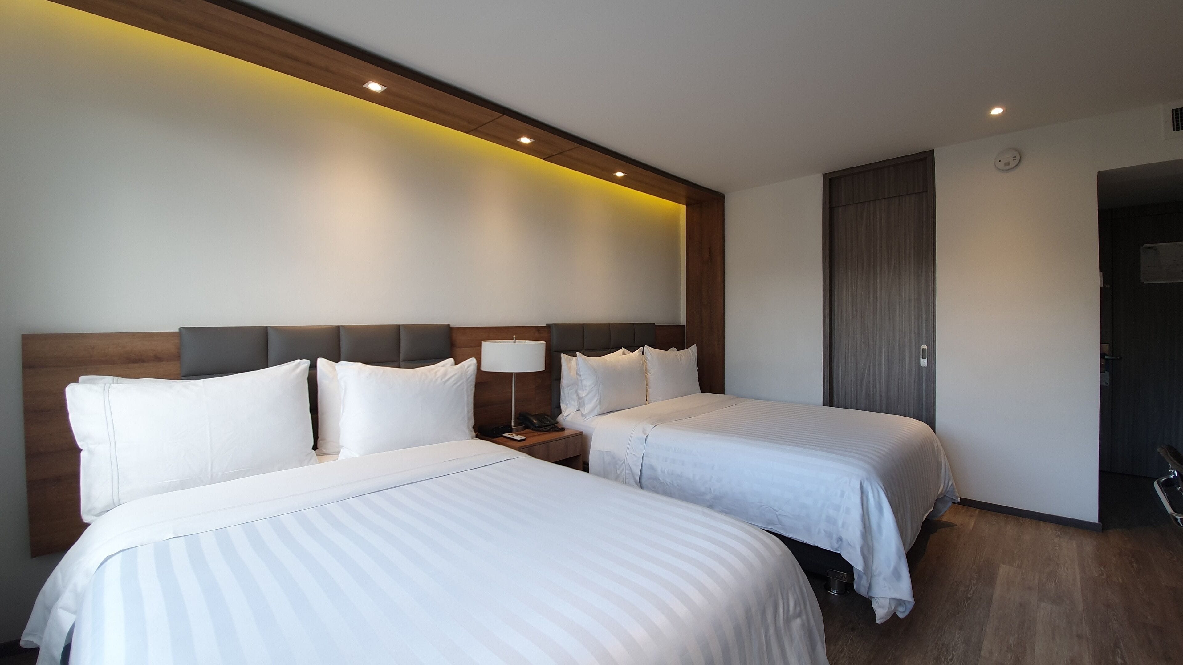 A hotel room with two beds, a bedside table with a lamp, and a door.