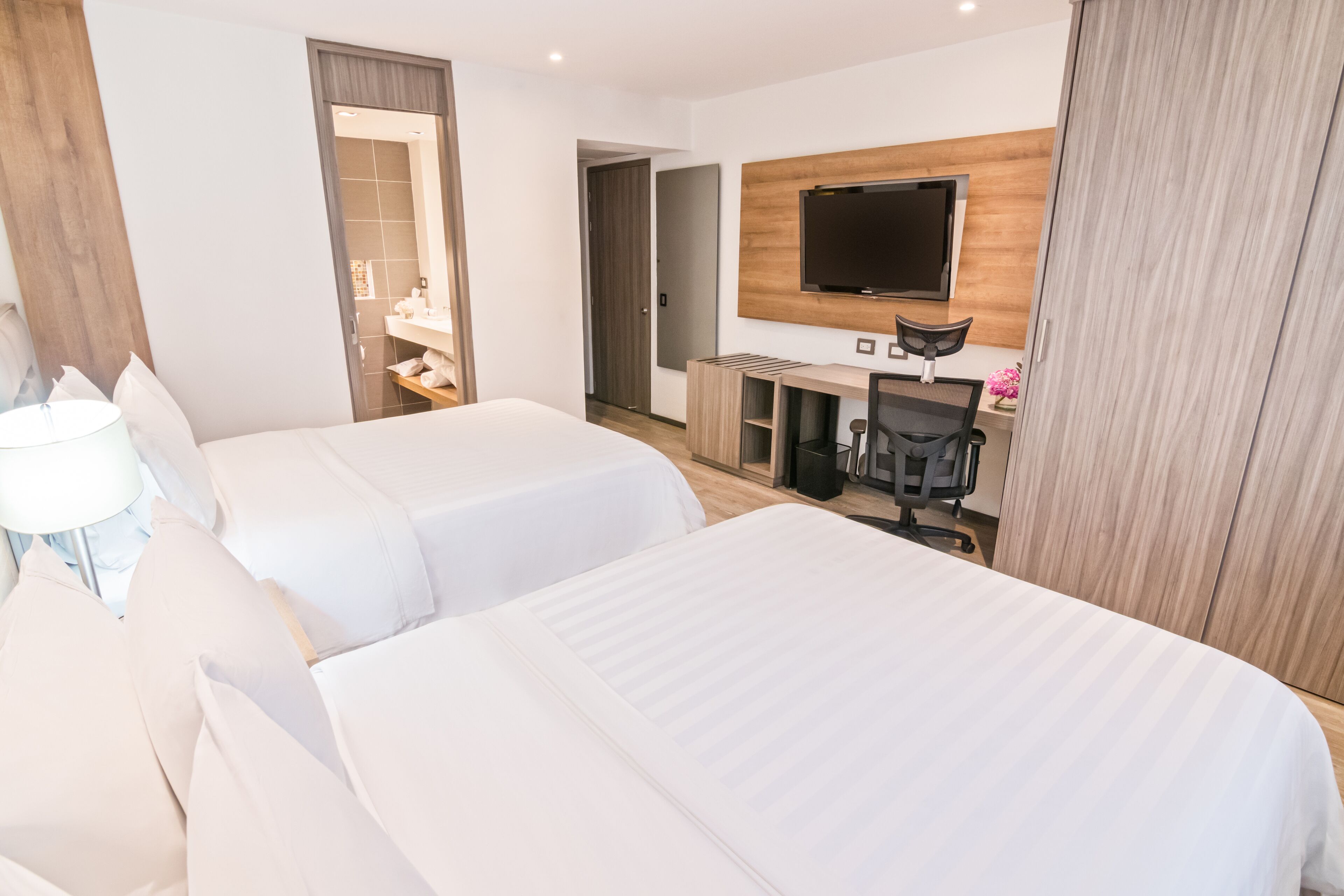 A hotel room with two beds, a desk with a chair, a TV mounted on the wall, and a wardrobe.