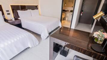 Habitación de hotel con dos camas, un escritorio con silla, una mesita con un florero y un baño visible a través de una puerta abierta.