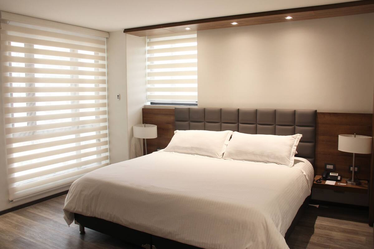 A modern hotel room with a large bed, two bedside tables with lamps, and a window with blinds.