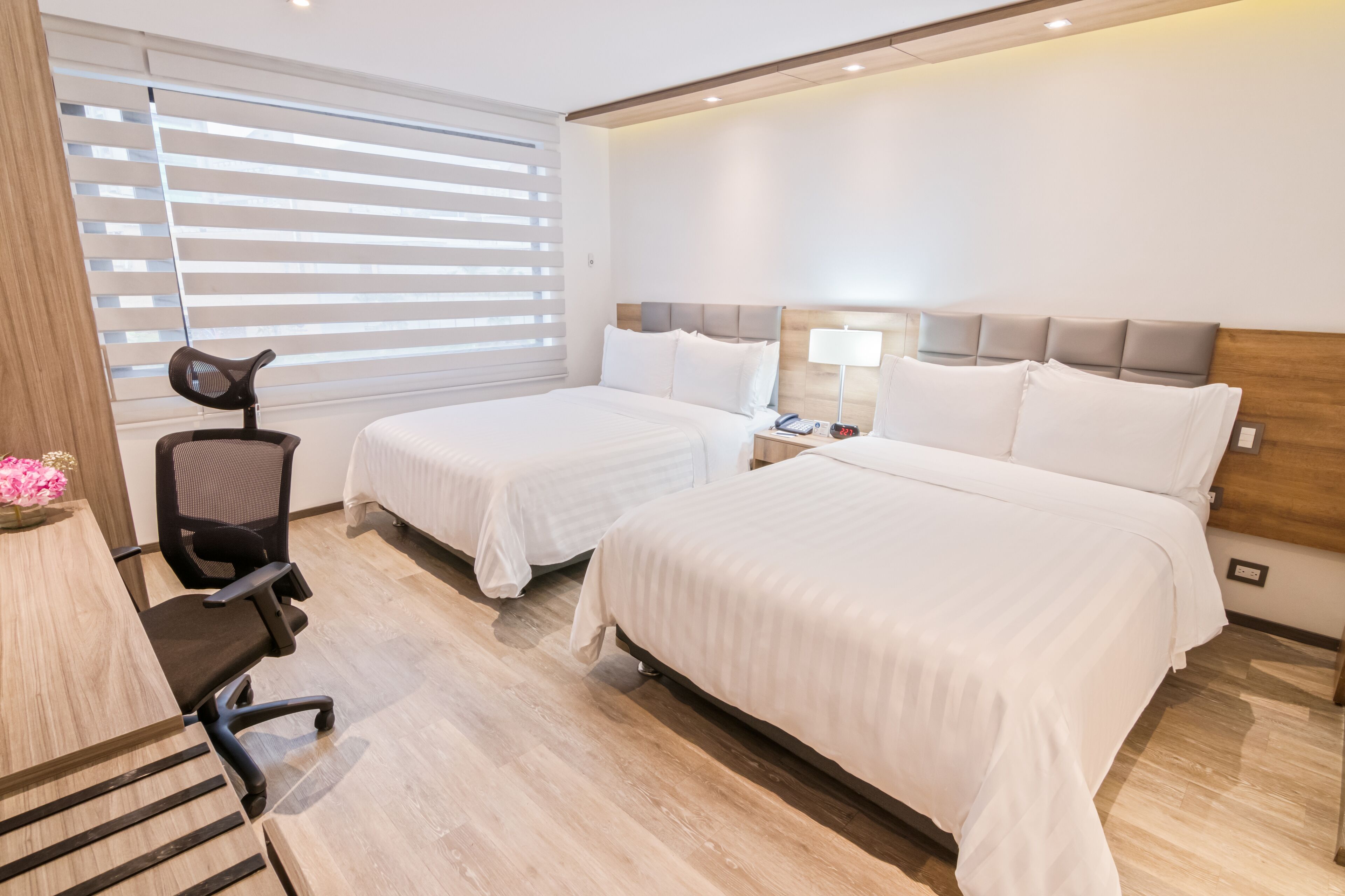 Habitación de hotel con dos camas, un escritorio con silla, una lámpara y una ventana con persianas.