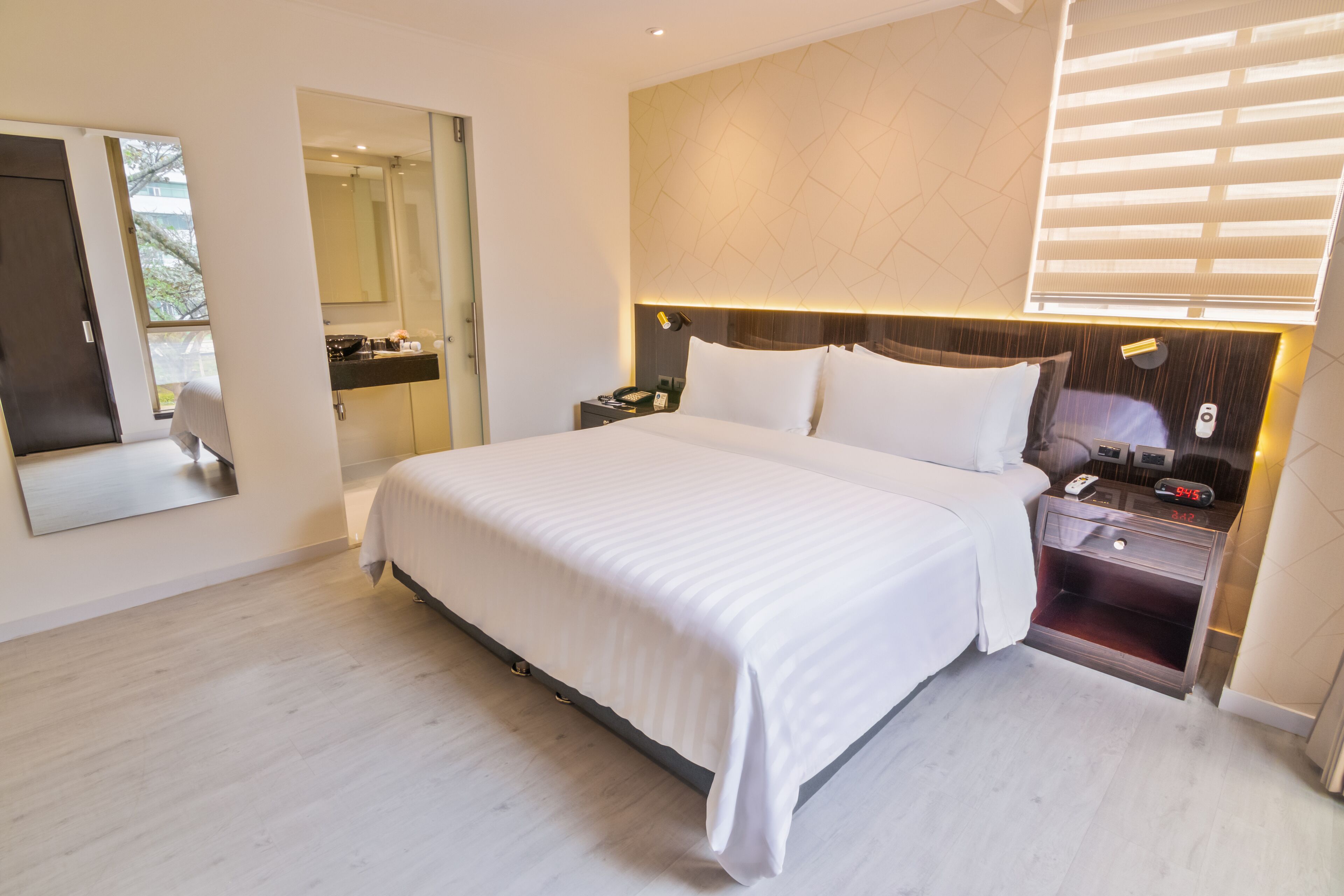 A modern hotel room with a large bed, a bedside table with a lamp, a mirror, and a nightstand with a phone and alarm clock.