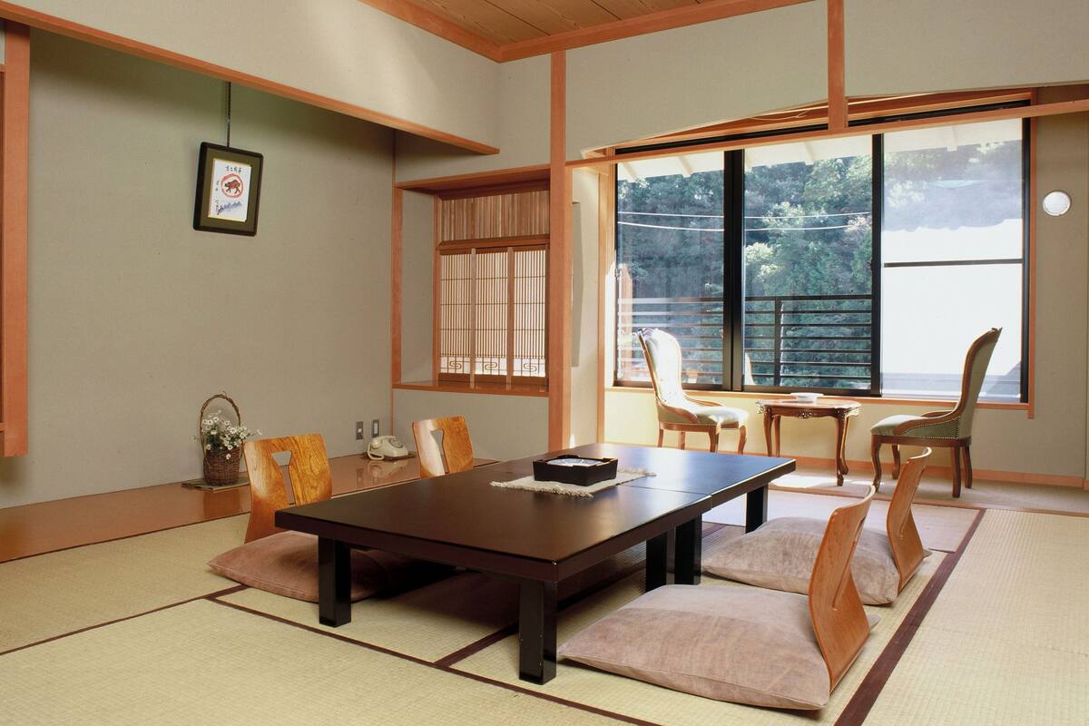 A traditional Japanese-style room with tatami flooring, a low wooden table, and a view of a forest through large windows.