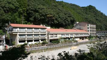 A multi-story hotel with a red roof, located near a beach with a rocky shoreline and dense forest in the background.