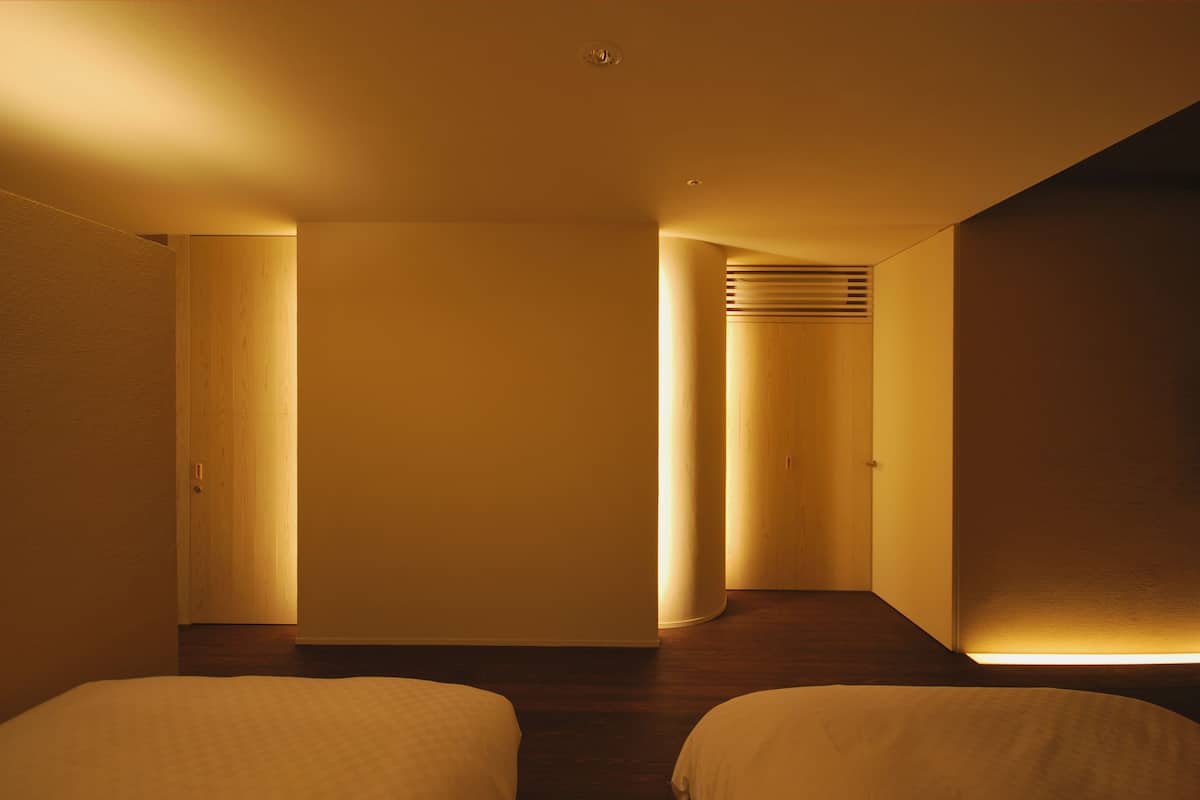 A hotel room with a wooden floor, two beds, a large window, and a door leading to another room.