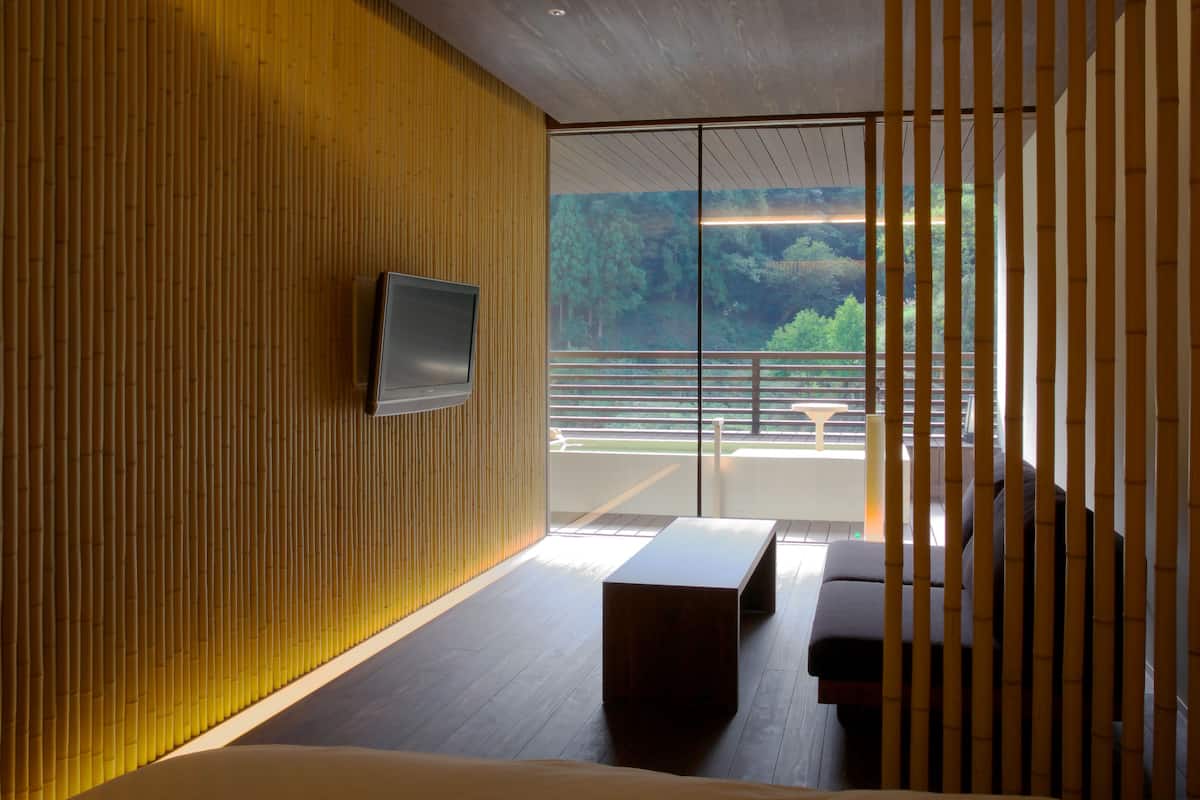 A modern room with a wooden wall, a flat-screen TV, a sofa, and a view of greenery through large windows.