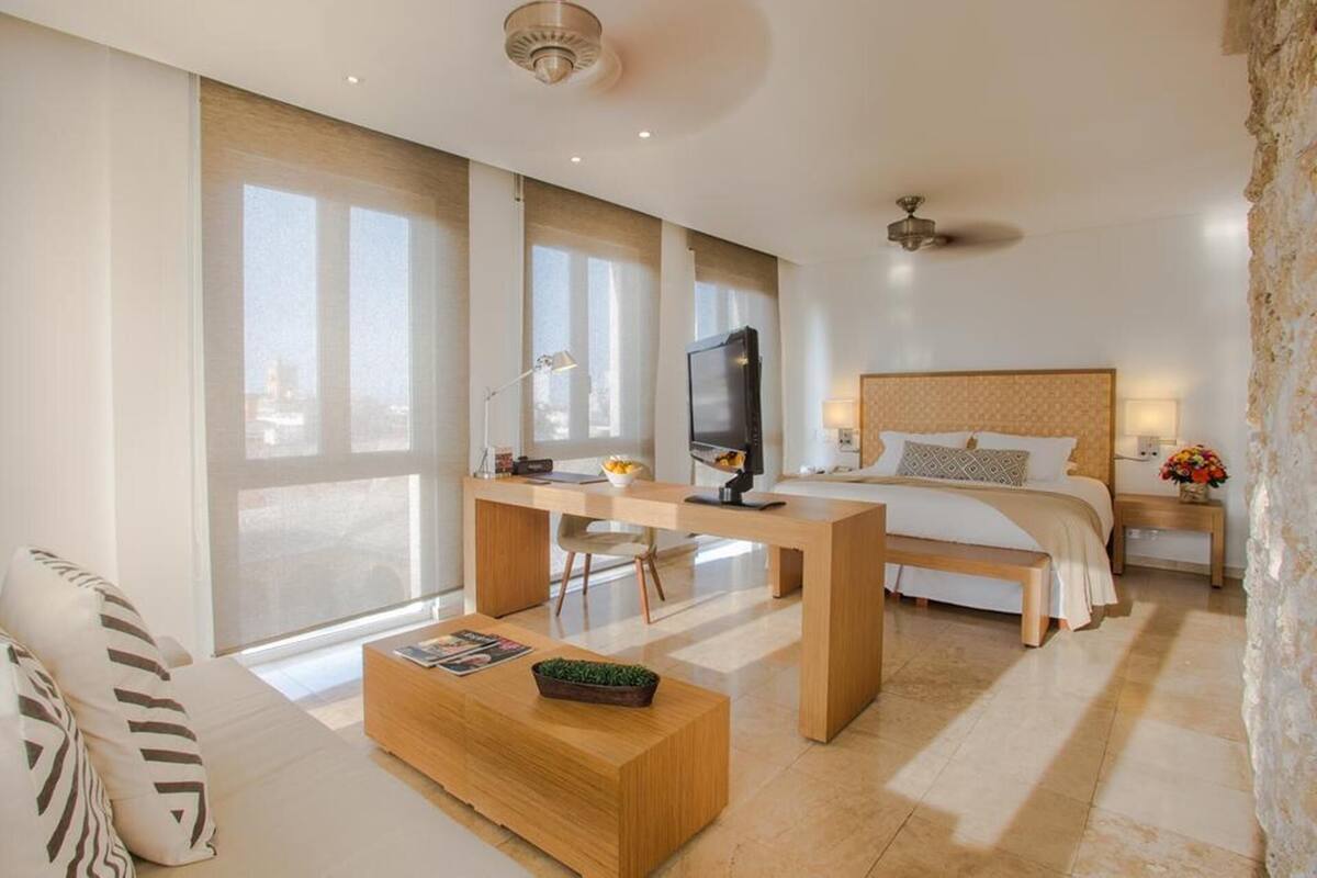 A modern hotel room with a large bed, a desk with a TV, a sofa, and a view of the city.