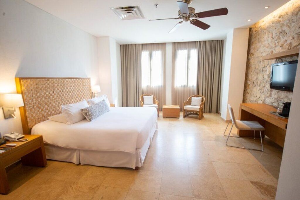 A hotel room with a large bed, a desk, a chair, a television, and a ceiling fan.