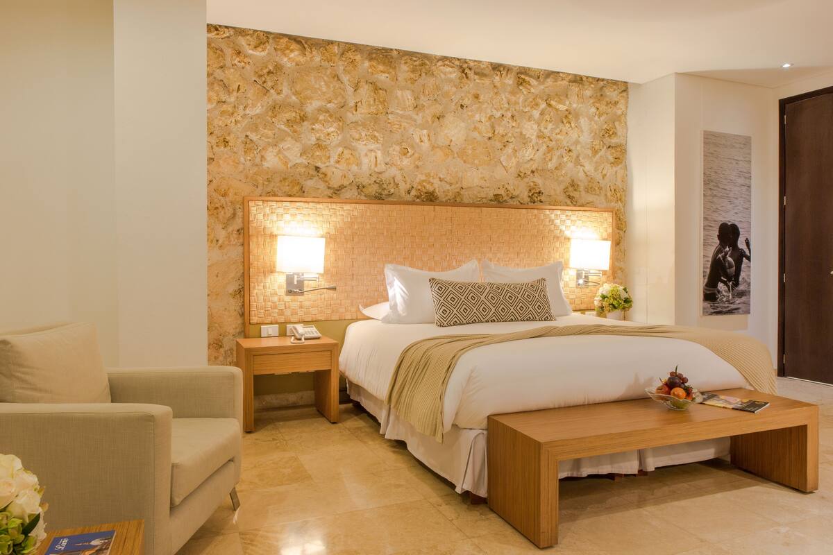A hotel room with a large bed, a bedside table with a lamp, a wooden bench, and a chair.