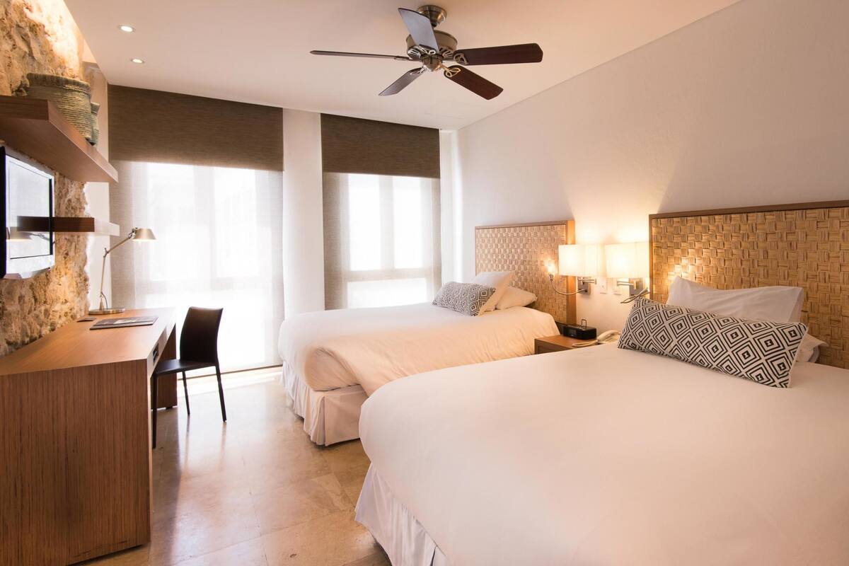 A hotel room with two beds, a desk, a chair, a ceiling fan, and a large window.