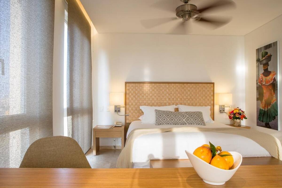 A hotel room with a bed, a desk, a chair, a ceiling fan, and a wall-mounted lamp.