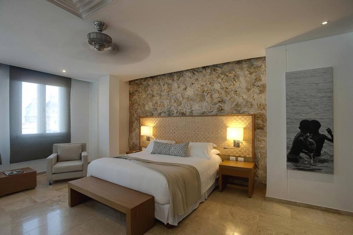 A bedroom with a large bed, two bedside lamps, a bench, a chair, a stone wall, and a framed picture.
