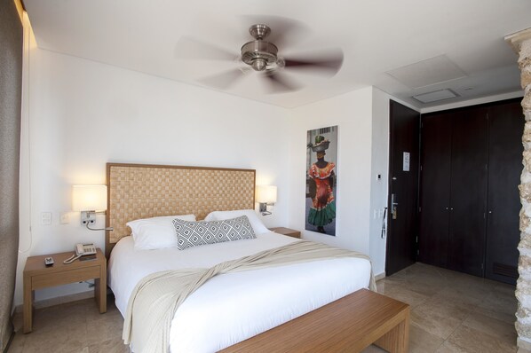 A hotel room with a large bed, a wooden headboard, two bedside lamps, a ceiling fan, a nightstand with a phone, a bench, and a wardrobe.