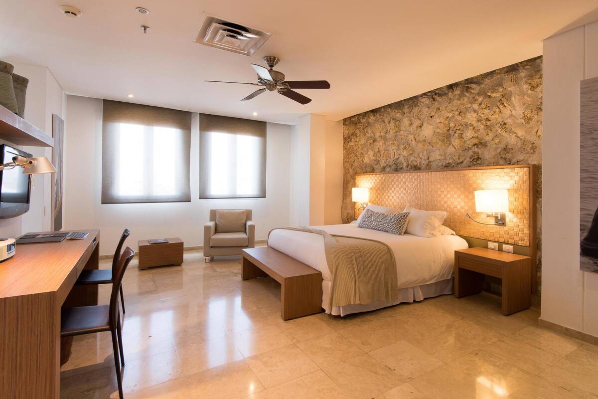 A modern hotel room with a large bed, a desk, a chair, a ceiling fan, and a stone-textured wall.