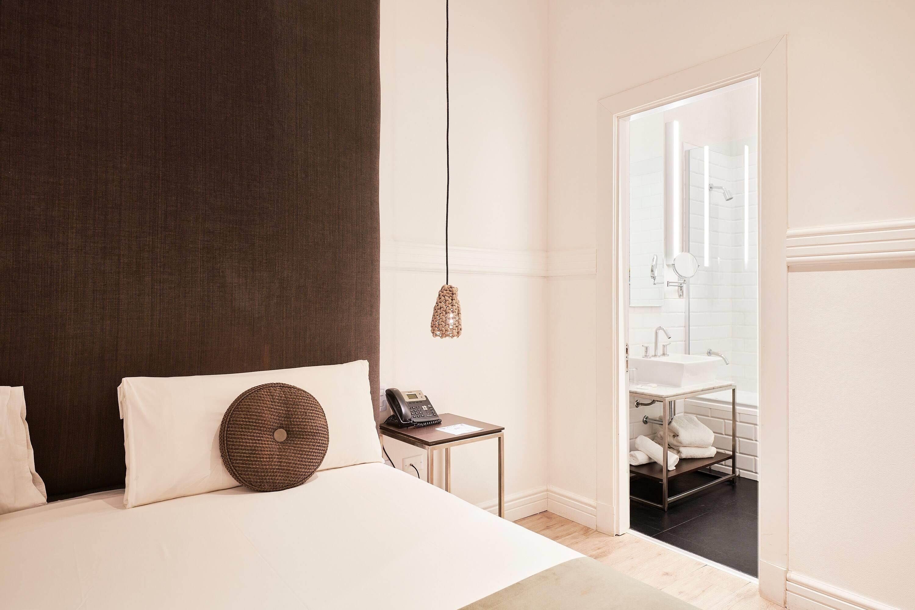 A hotel room with a bed, bedside table with a phone, a hanging lamp, and a view of the bathroom.