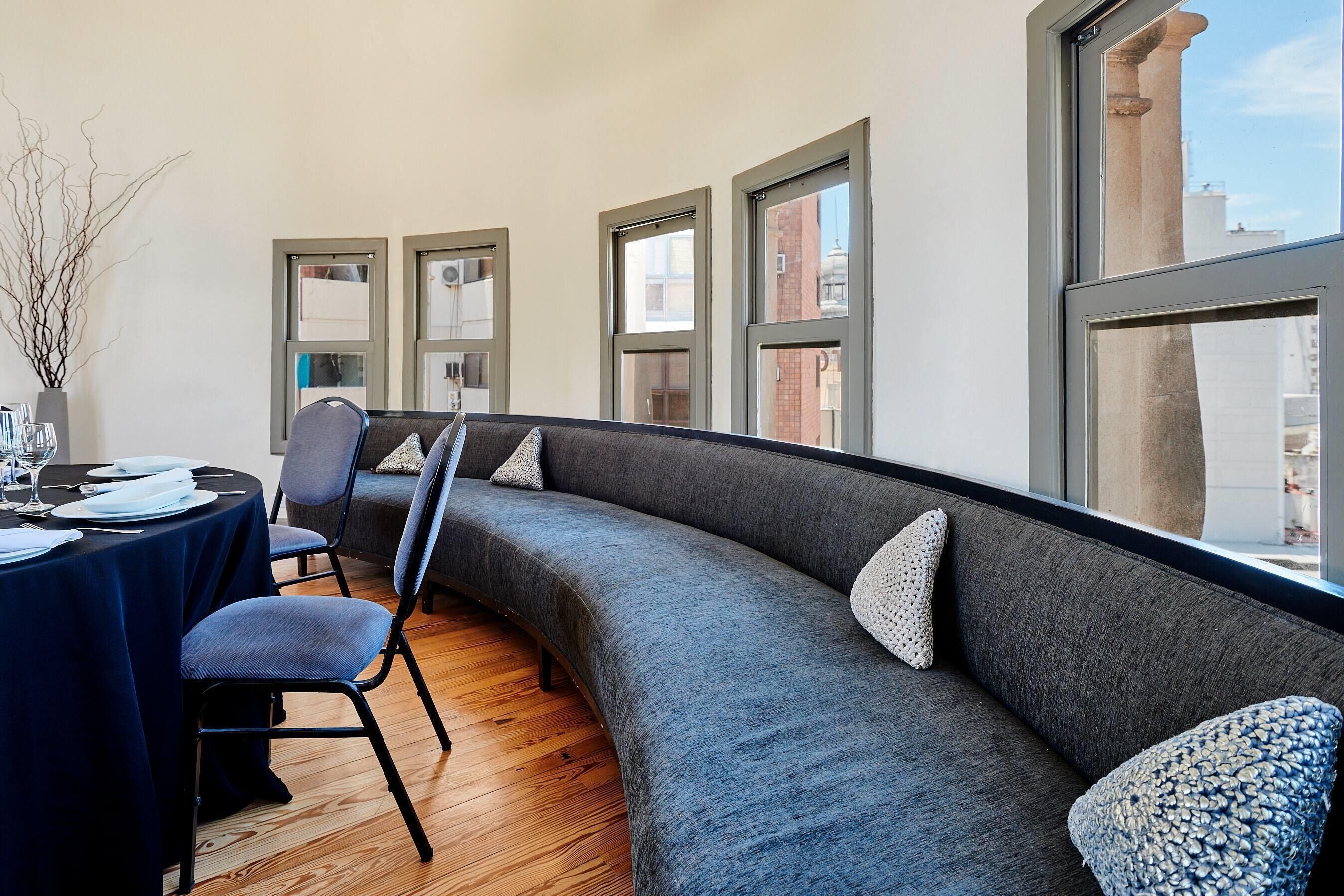 A modern room with a curved gray sofa, a dining table set with plates and glasses, and large windows offering a view of the city.