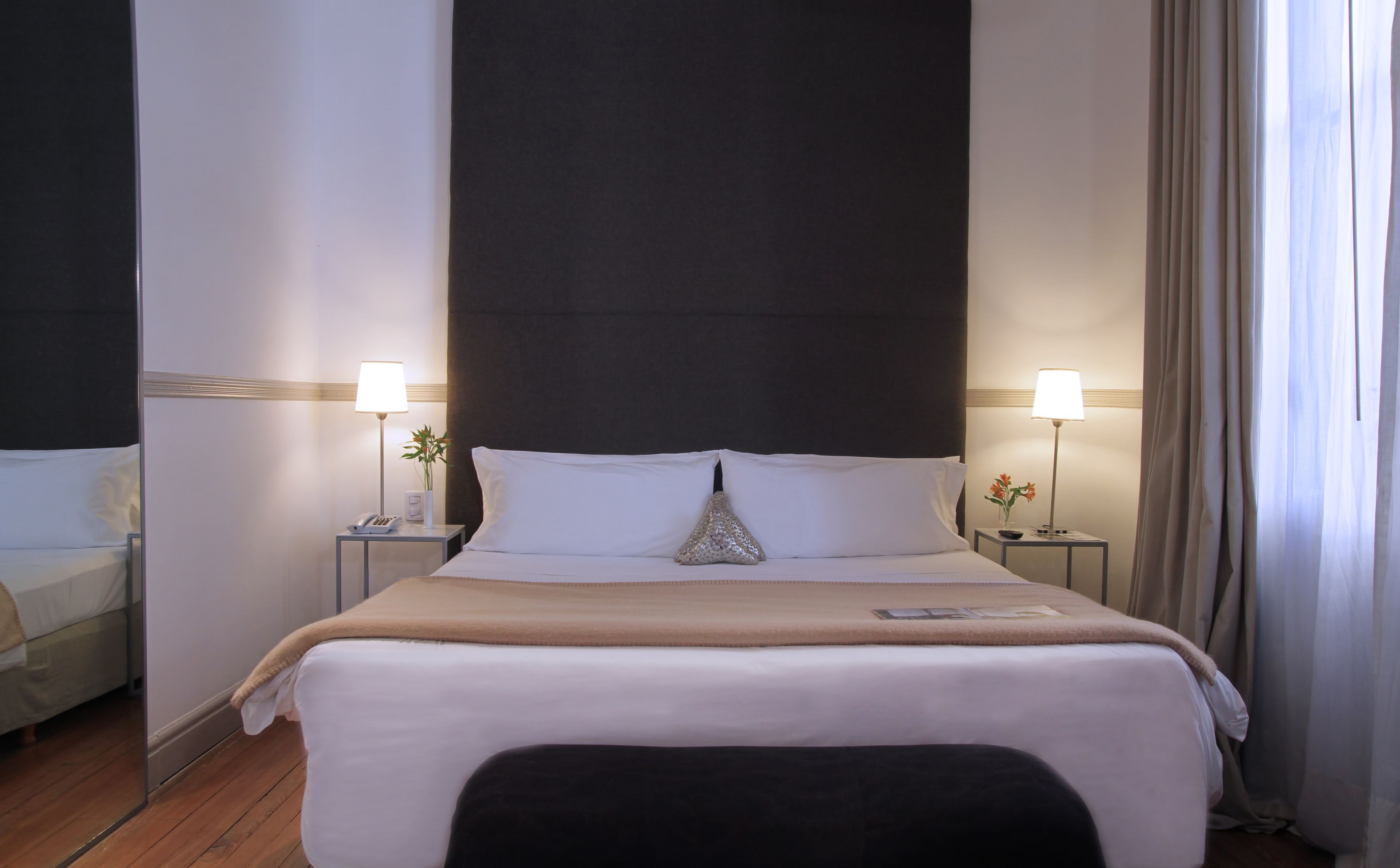 A hotel room with a large bed, two bedside tables with lamps, a small vase with flowers, and a window with curtains.