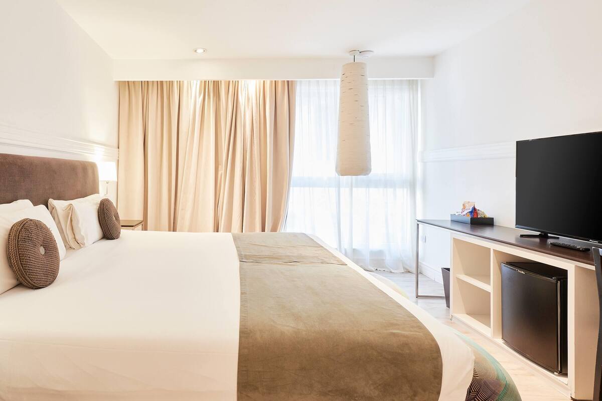 A hotel room with a large bed, a flat-screen TV, a small refrigerator, and a window with sheer curtains.