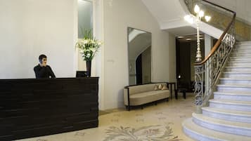 Zona de recepción con un mostrador oscuro, un sofá, una escalera con barandillas decoradas y una alfombra estampada con motivos florales.