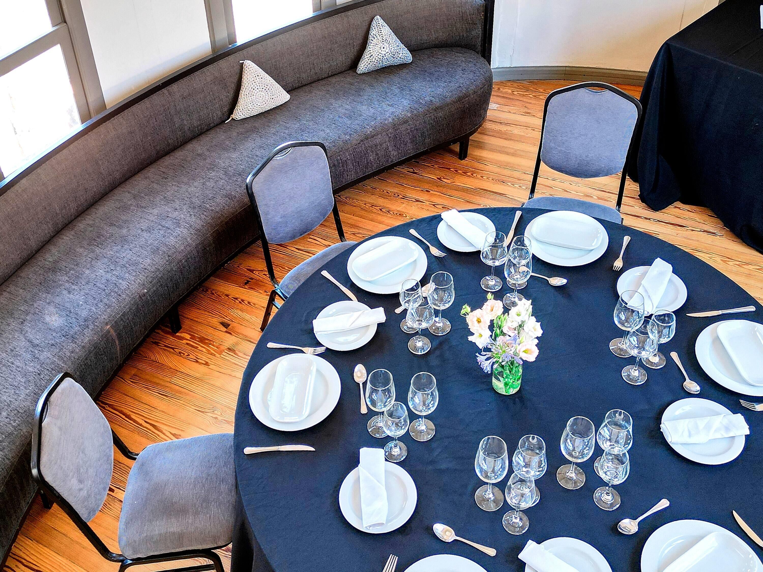A round dining table set with white plates, glasses, and cutlery, surrounded by grey upholstered chairs, with a grey sofa and pillows in the background.