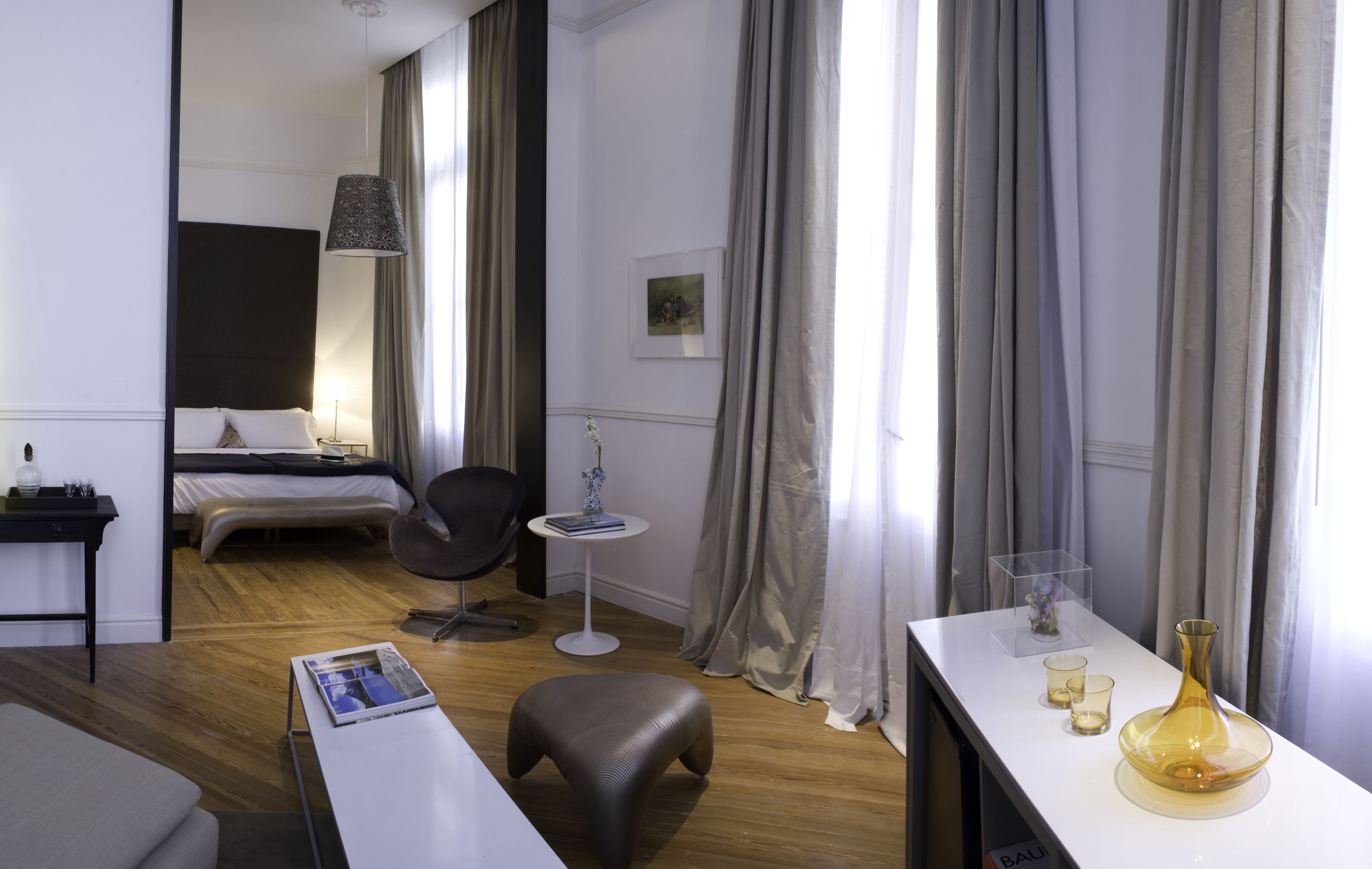 A hotel room with a bed, bedside table, chair, small table with a vase, and a window with curtains.