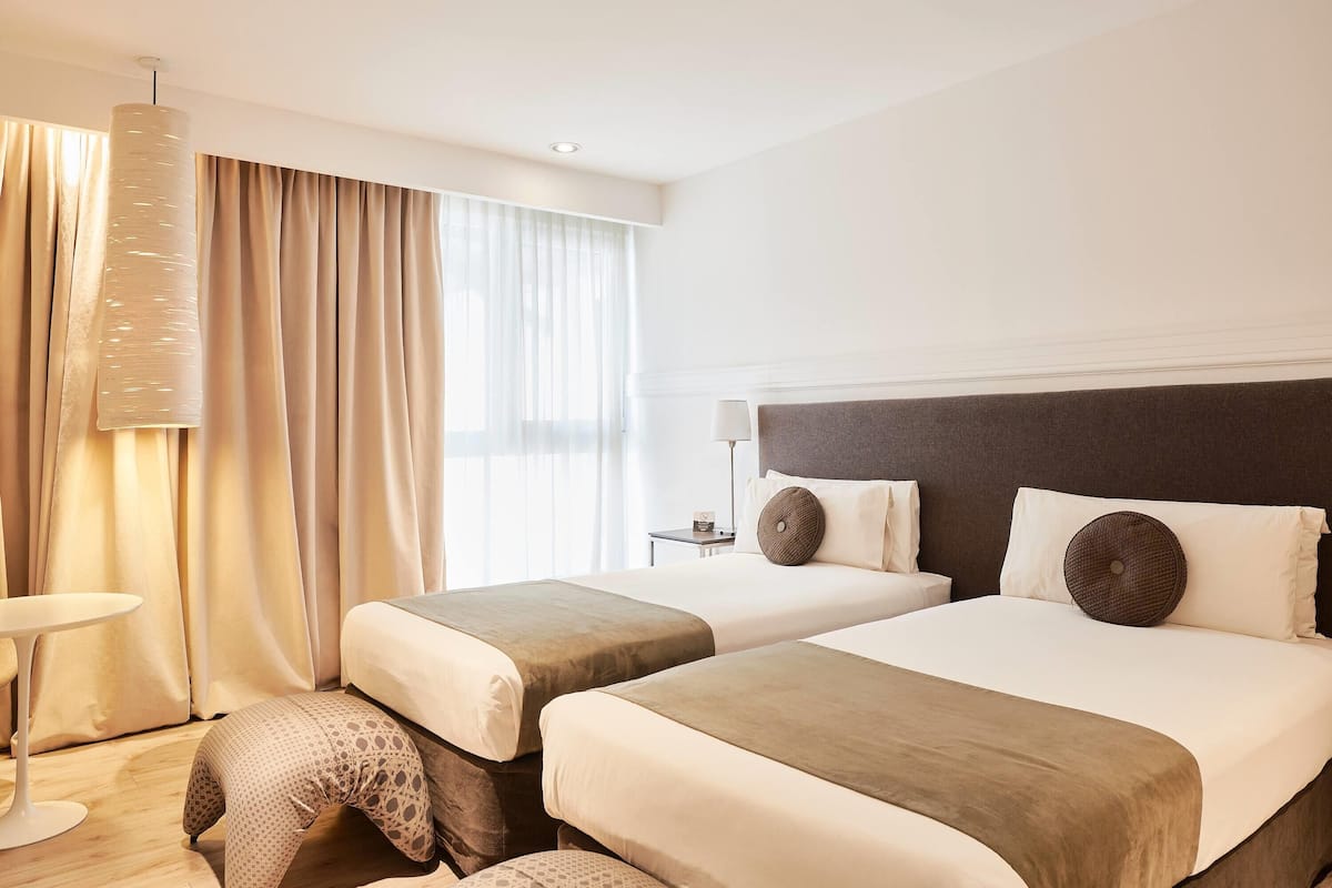 A hotel room with two beds, a bedside table, a lamp, a hanging light fixture, and a window with curtains.