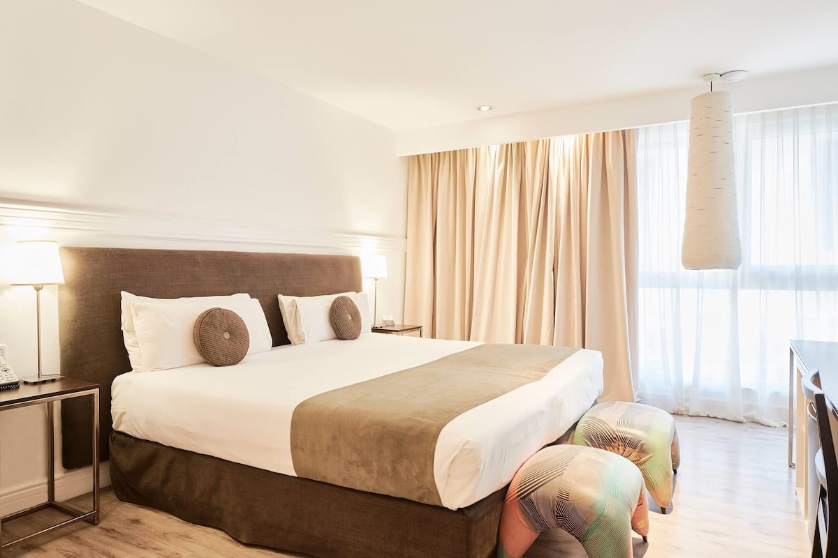 A hotel room with a large bed, a nightstand with a lamp, and a view of the outdoors through sheer curtains.