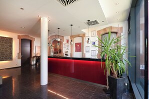 A modern hotel lobby with a red reception desk, a large column, and a potted plant. - NOVINA HOTEL Tillypark (Nuremberg)