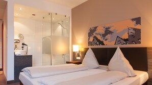 A hotel room with a large bed, a shower, a vanity with a mirror, and a painting on the wall. - NOVINA HOTEL Tillypark (Nuremberg)