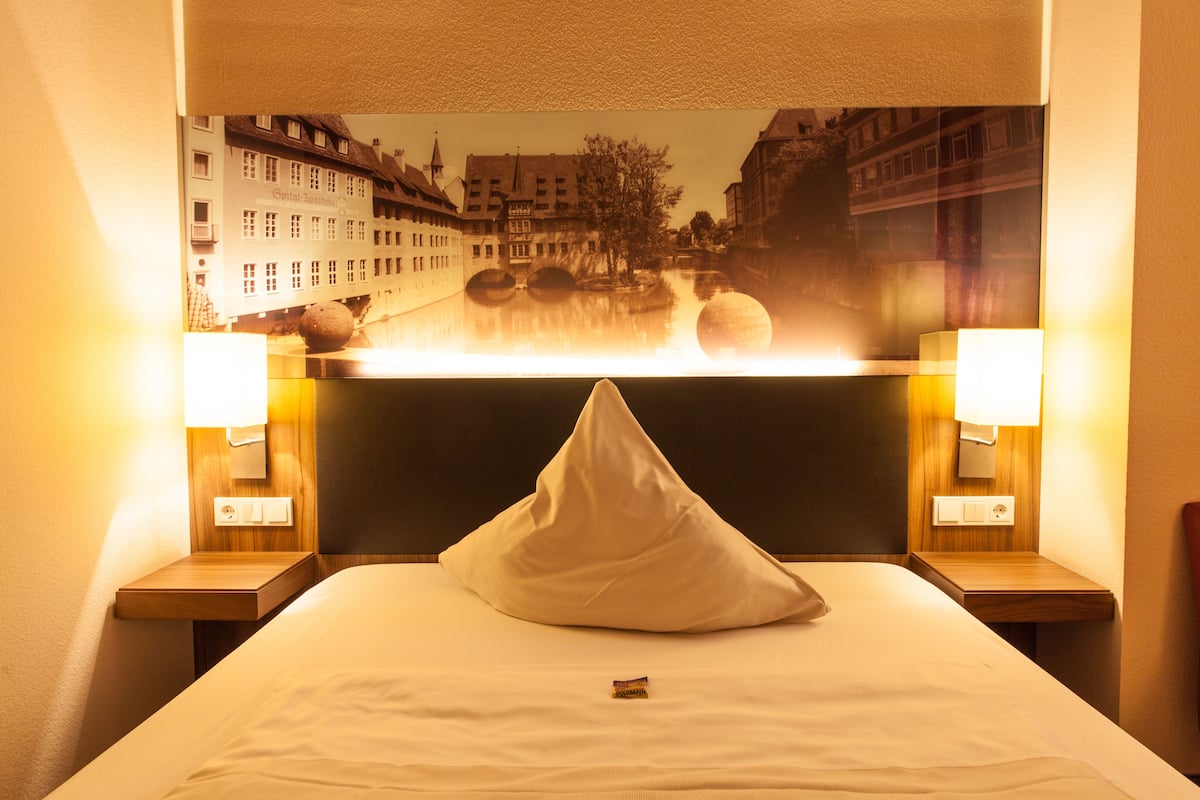 A hotel room with a bed, a pillow, two bedside lamps, and a wall-mounted picture of a cityscape.