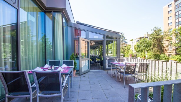 An outdoor patio with tables and chairs, a glass enclosure, and a view of the surrounding buildings and greenery. - NOVINA HOTEL Tillypark (Nuremberg)