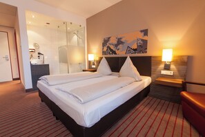 A hotel room with a large bed, a shower area, a desk with a mirror, and two bedside lamps. - NOVINA HOTEL Tillypark (Nuremberg)