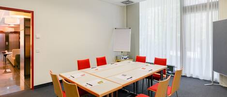 Une salle de conférence avec une longue table, des chaises rouges, un tableau blanc et une grande fenêtre avec des rideaux.