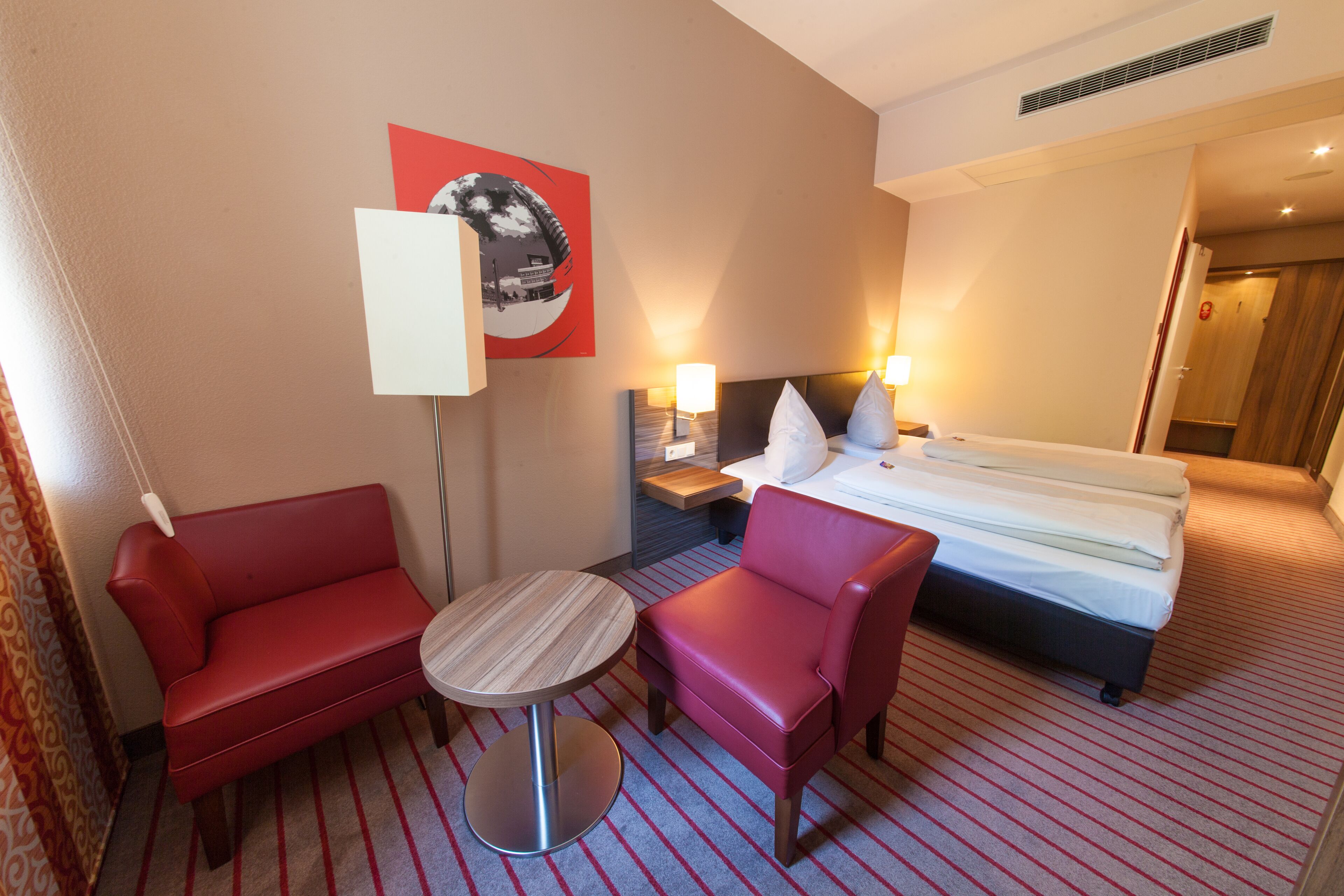 A hotel room with a bed, two red sofas, a small round table, and a wall-mounted TV.
