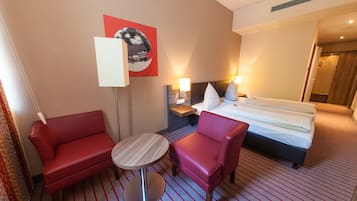 Habitación de hotel con una cama, dos sofás rojos, una mesita redonda y un televisor instalado en la pared.